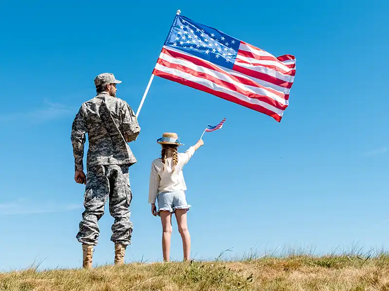 Service member and child holding American flags on grassy hill