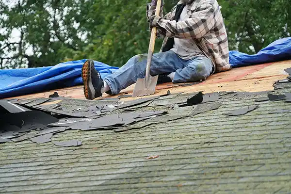 Roofing worker removing old shingles from roof with shovel